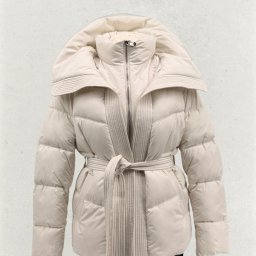 KM INVESTMENT POLAND PATRYK MIŁOSZ - Beżowa, pikowana kurtka puchowa z szerokim kołnierzem i wiązanym paskiem, na neutralnym tle. Widoczny fragment czarnych spodni. Elegancki krój, ciepła odzież wierzchnia.