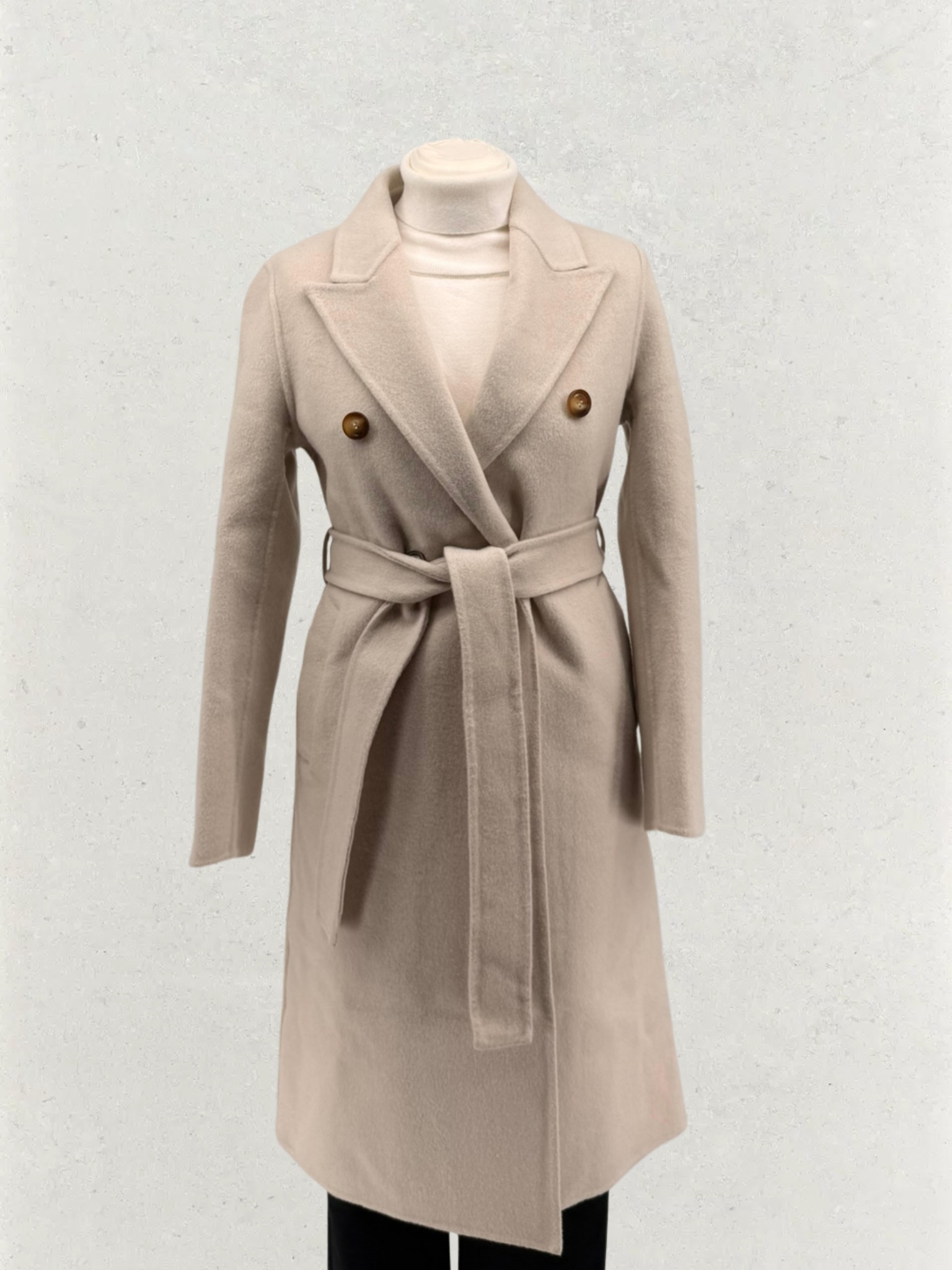 Beżowy, wełniany płaszcz damski z paskiem, zapinany na dwa guziki, prezentowany na manekinie na tle jasnej ściany. Elegancki krój, idealny na chłodniejsze dni.