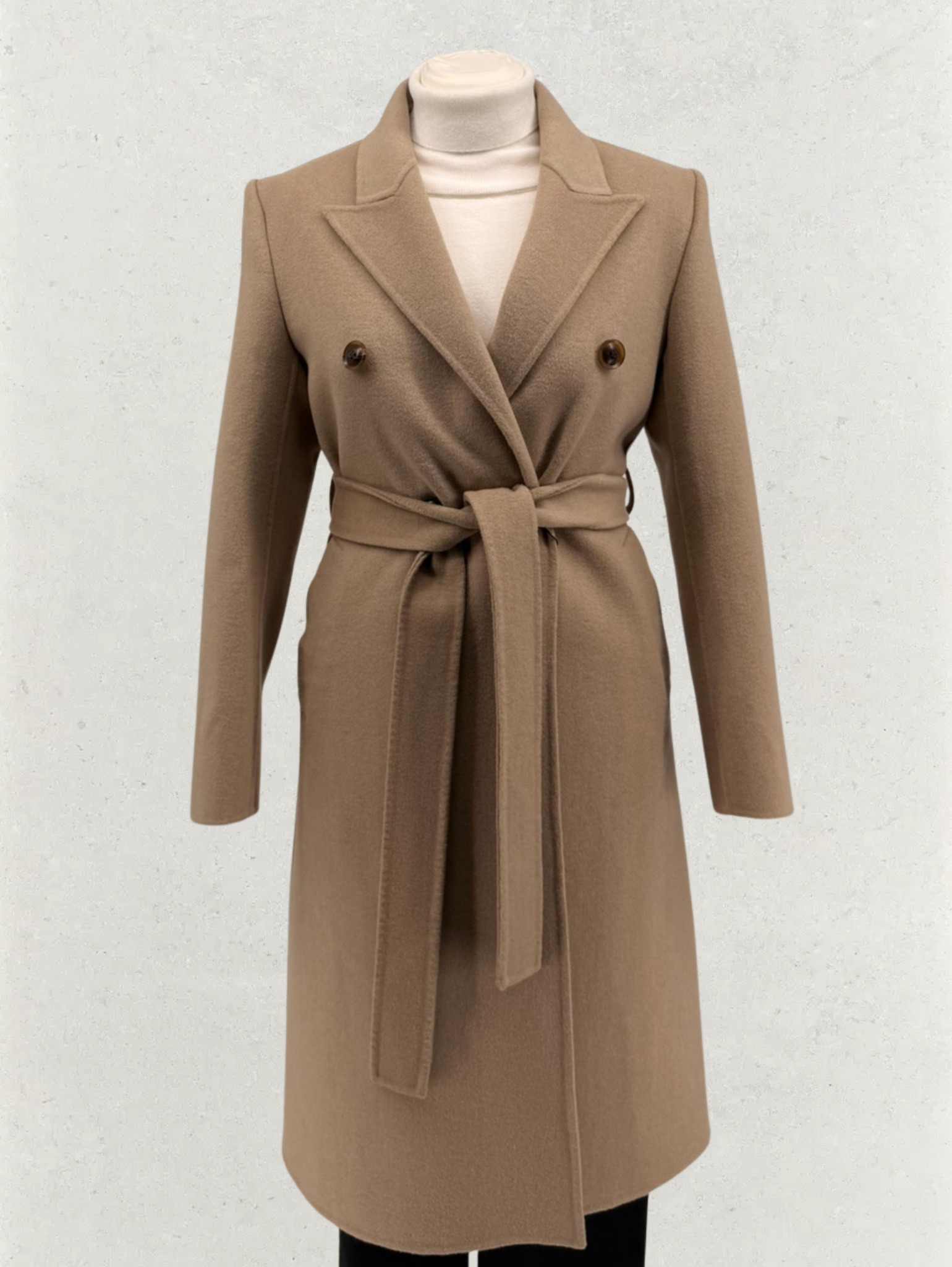 Beżowy, wełniany płaszcz damski z paskiem, dwurzędowy, na manekinie. Widoczne detale: guziki, klapy, wiązanie w pasie. Stonowana kolorystyka, gładka faktura materiału.