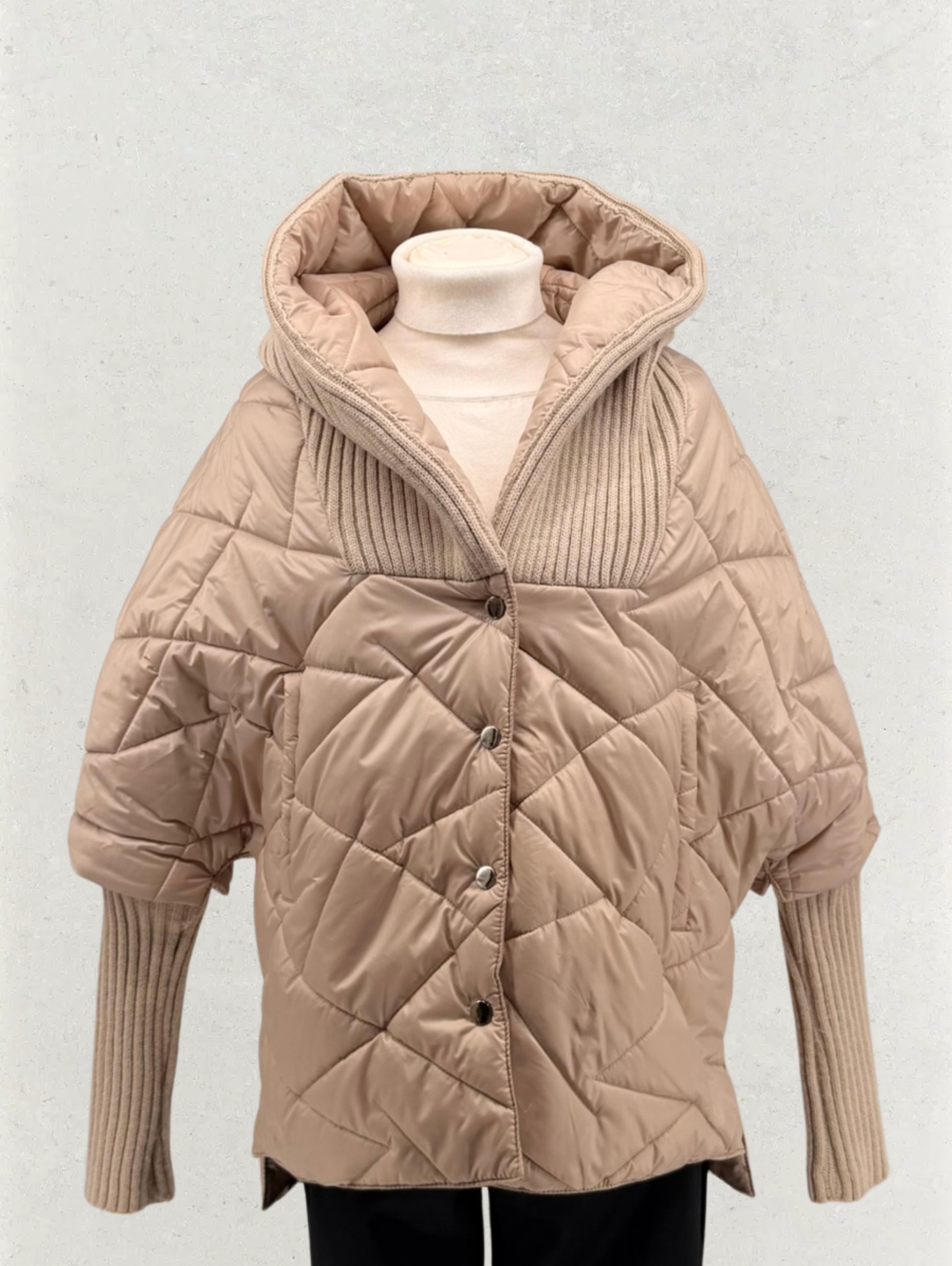 Beżowa, pikowana kurtka z kapturem i dzianinowymi rękawami, zapinana na guziki, na manekinie. Widoczne detale: faktura pikowania, splot dzianiny.