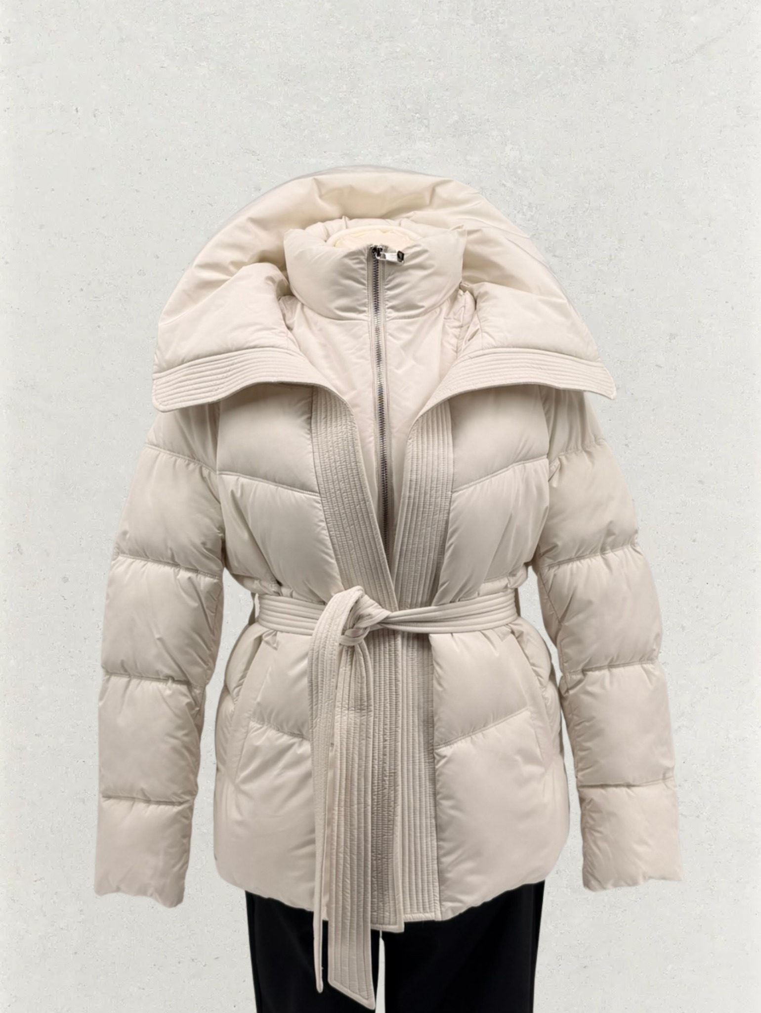 Beżowa, pikowana kurtka puchowa z szerokim kołnierzem i wiązanym paskiem, na neutralnym tle. Widoczny fragment czarnych spodni. Elegancki krój, ciepła odzież wierzchnia.