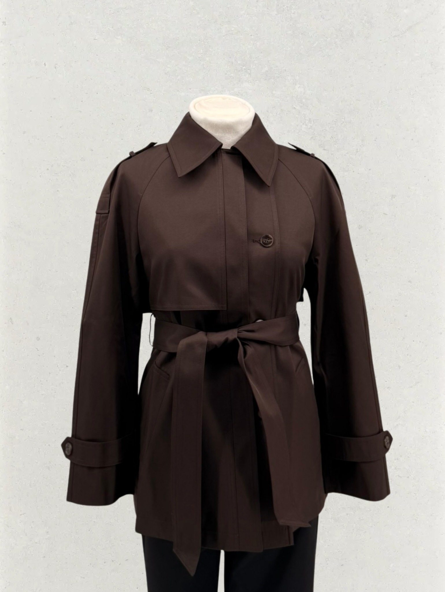 Brązowy trencz damski z paskiem, zaprezentowany na manekinie na tle jasnej ściany. Widoczne detale: kołnierz, pagony, mankiety z guzikami. Elegancki krój, gładki materiał.