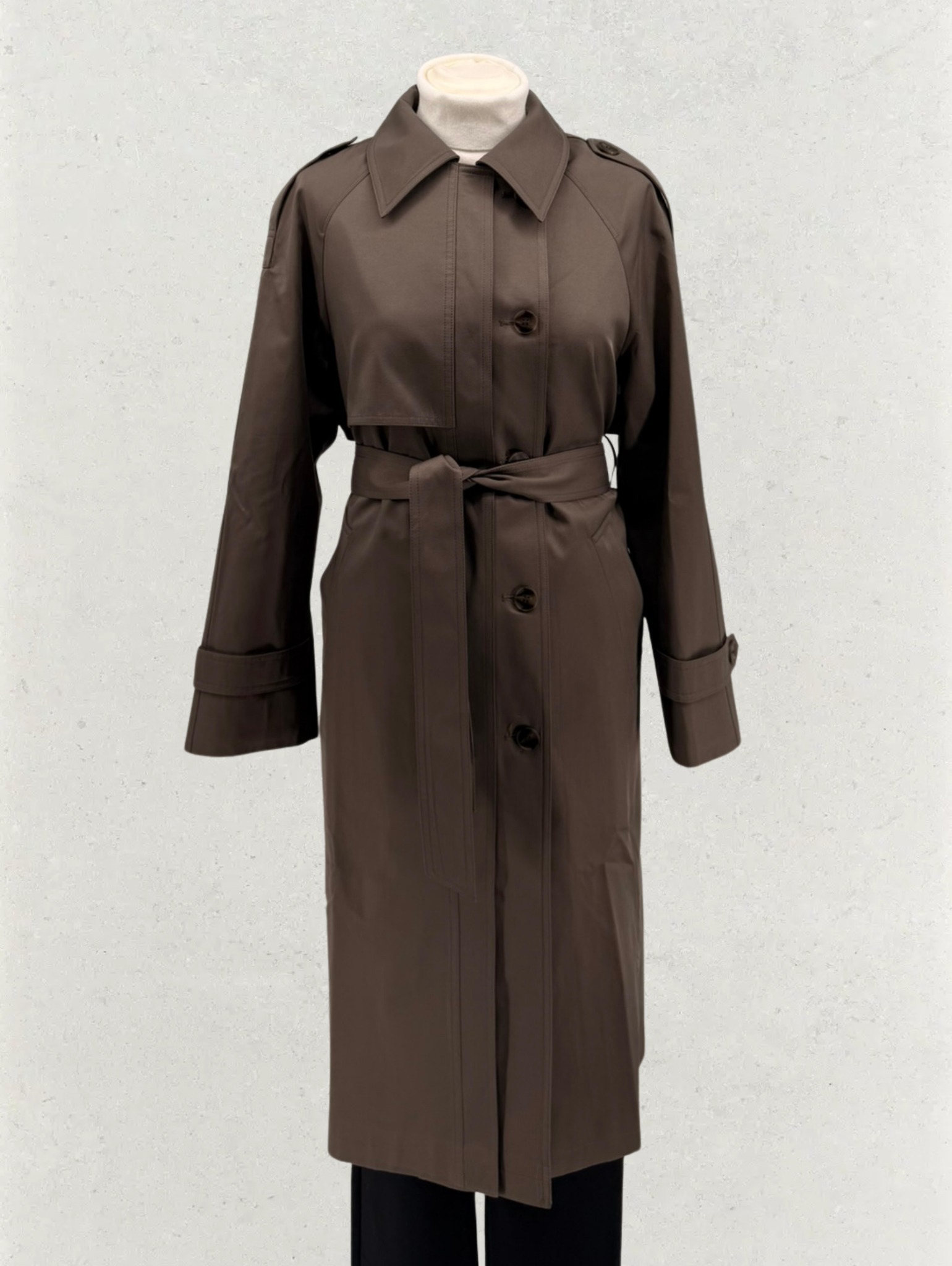 Brązowy, elegancki płaszcz damski z paskiem, zapinany na guziki, na manekinie. Stonowana kolorystyka, gładki materiał, klasyczny krój. Neutralne tło.