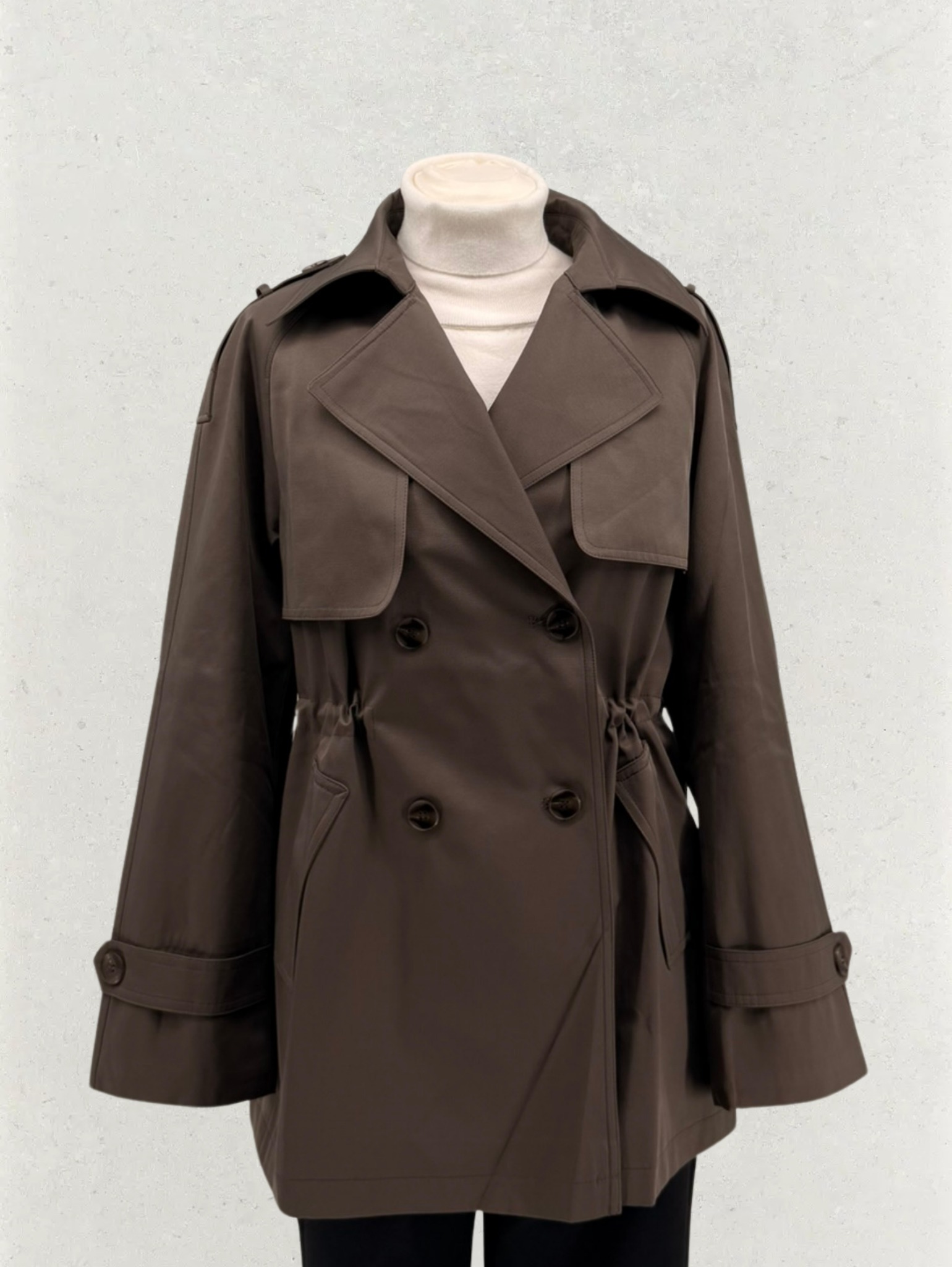 Brązowy, dwurzędowy płaszcz damski z wiązaniem w pasie, zaprezentowany na manekinie na tle jasnej ściany. Widoczne detale: klapy, guziki i regulowane mankiety.