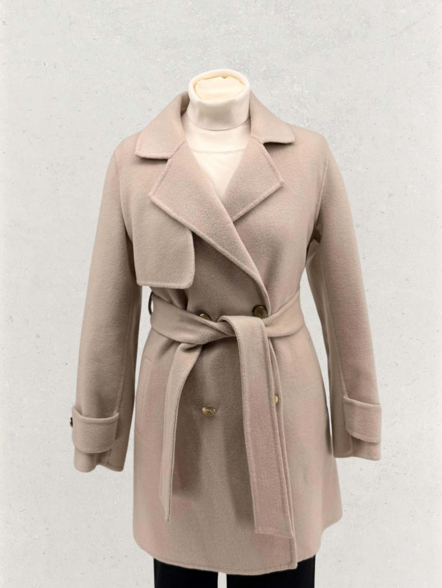 Beżowy, wełniany płaszcz damski z paskiem, zapinany na dwa guziki, prezentowany na manekinie na tle jasnej ściany. Widoczny kołnierz i mankiety.