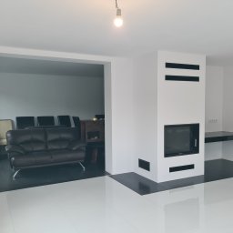 UMBRELLA-BUD DOMINIK SIERPIEŃ - Białe wnętrze z kominkiem i czarnymi akcentami: podłoga, półka, detale kominka. W tle widoczny salon z ciemną sofą. Minimalistyczny design, surowe wykończenie.