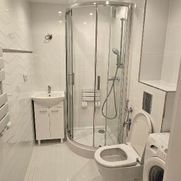 UMBRELLA-BUD DOMINIK SIERPIEŃ - Biała łazienka po remoncie: kabina prysznicowa, umywalka z szafką, grzejnik drabinkowy i pralka. Ściany wyłożone białymi płytkami z delikatnym wzorem.