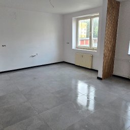 UMBRELLA-BUD DOMINIK SIERPIEŃ - Wykończone wnętrze: szare płytki podłogowe, białe ściany z drewnianymi panelami dekoracyjnymi i grzejnikami pod oknami. Widok na zieleń za oknem.