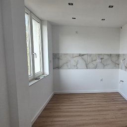 UMBRELLA-BUD DOMINIK SIERPIEŃ - Jasna kuchnia po remoncie: marmurowe płytki nad blatem, drewniana podłoga, białe ściany i okno z widokiem na zieleń. Minimalistyczne oświetlenie sufitowe.