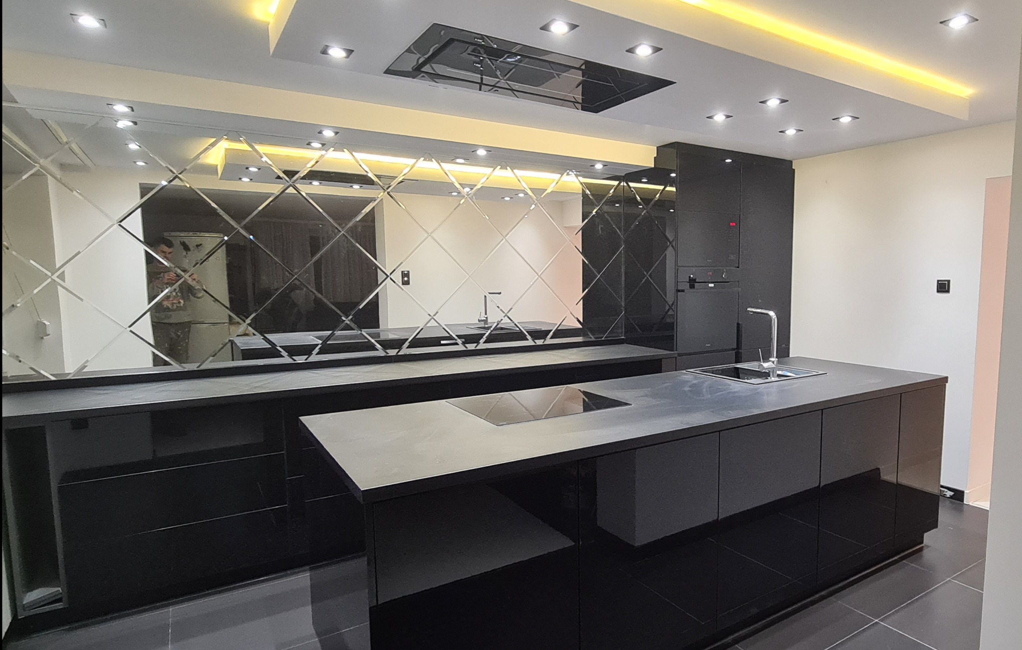 Nowoczesna, czarna kuchnia z wyspą i lustrzaną ścianą w romby. Oświetlenie sufitowe podkreśla minimalistyczny design. Widoczny fragment podłogi z ciemnych płytek.