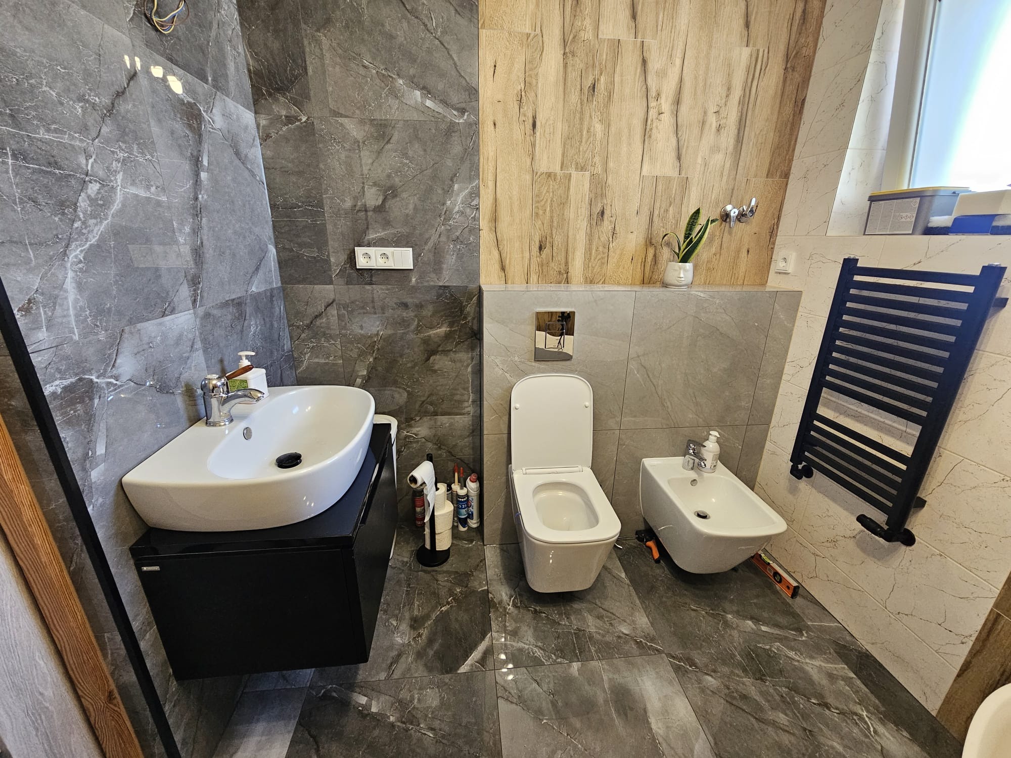 Nowoczesna łazienka po remoncie: marmurowe płytki, drewniane akcenty, biała armatura (umywalka, toaleta, bidet), czarny grzejnik drabinkowy. Widok na całe pomieszczenie.