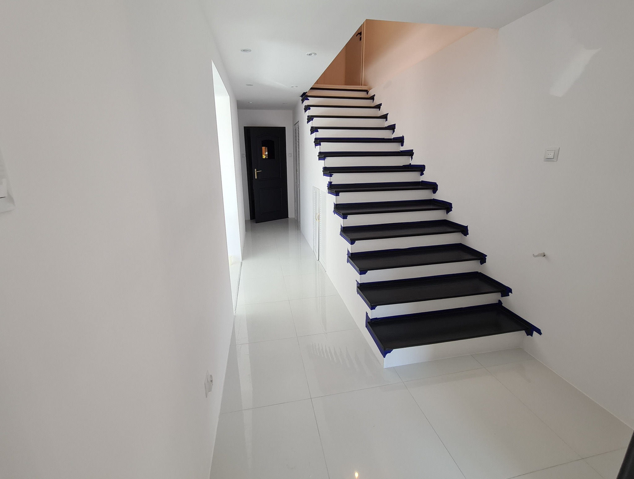 Białe wnętrze po malowaniu: schody z czarnymi stopniami zabezpieczone taśmą malarską, białe ściany i podłoga z połyskiem, czarne drzwi w tle.