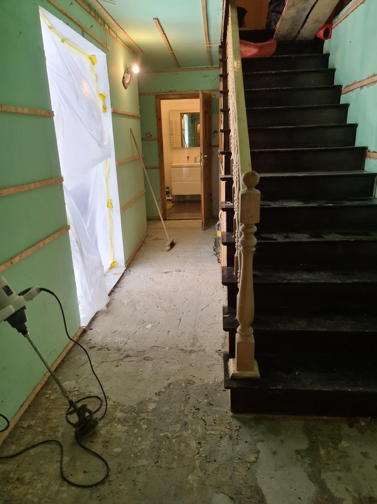Remont klatki schodowej w trakcie: zabezpieczone folią drzwi, ściany z płyt karton-gips, schody drewniane. Widoczny mikser do zaprawy i fragment łazienki w tle.