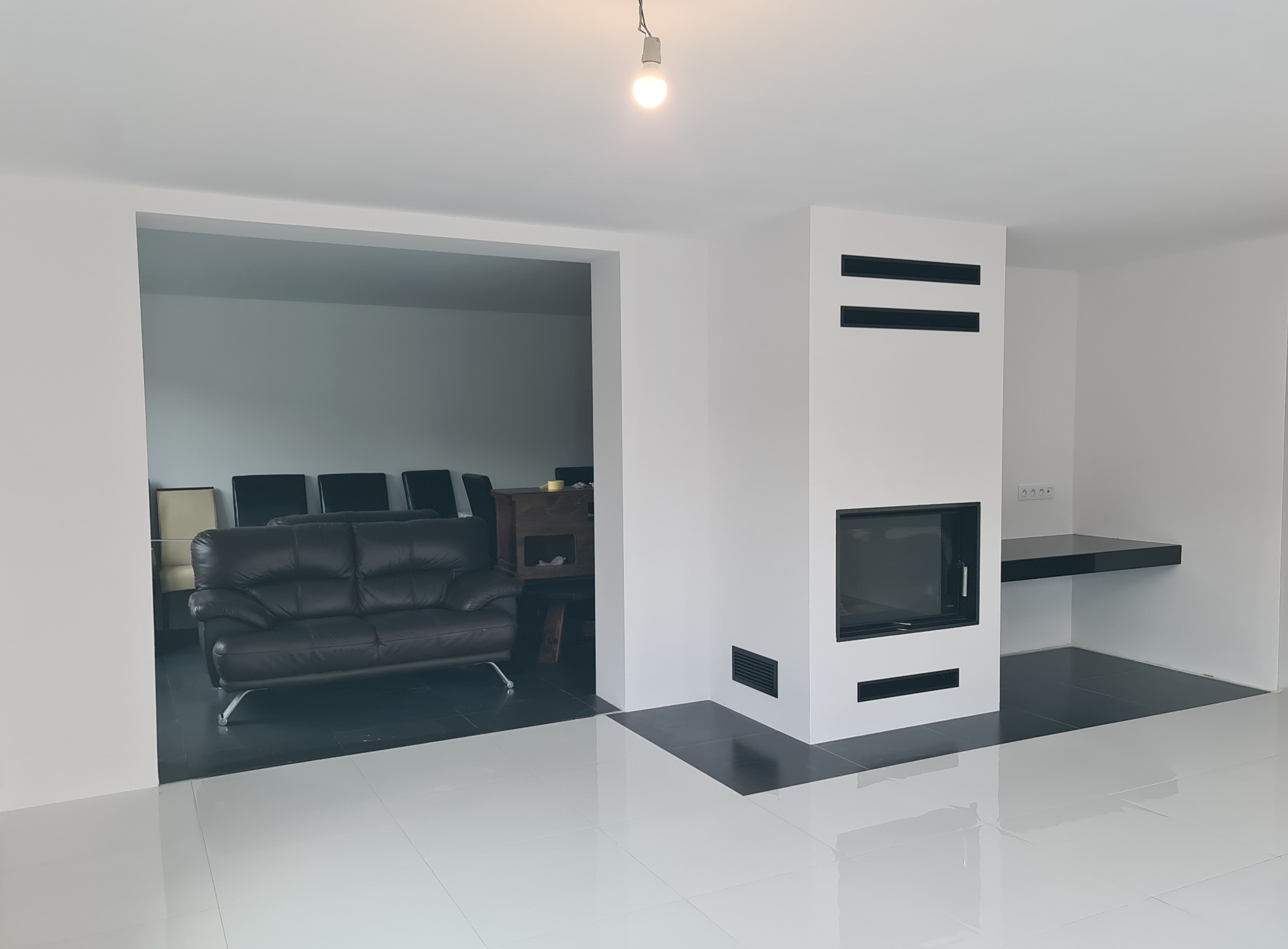 Białe wnętrze z kominkiem i czarnymi akcentami: podłoga, półka, detale kominka. W tle widoczny salon z ciemną sofą. Minimalistyczny design, surowe wykończenie.