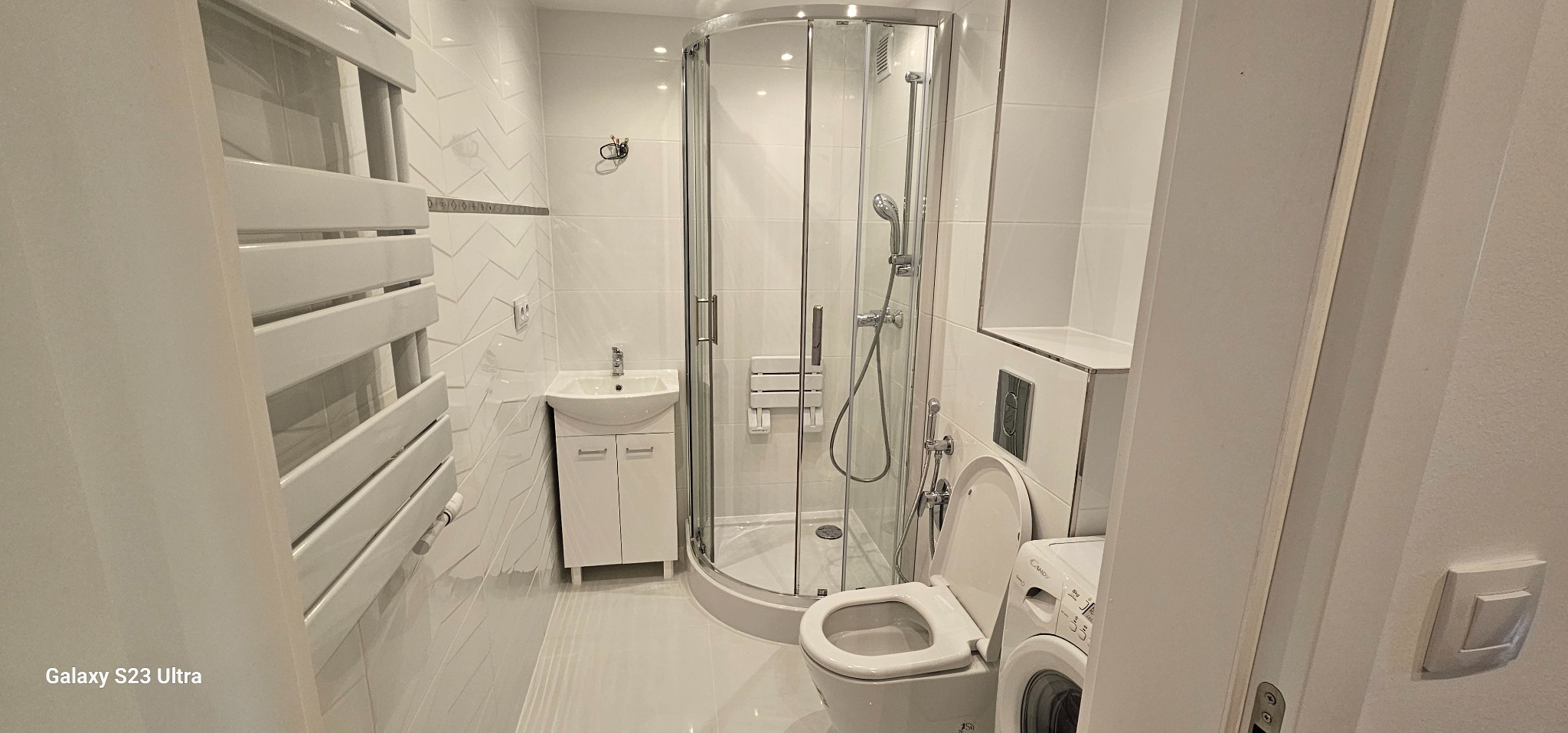 Biała łazienka po remoncie: kabina prysznicowa, umywalka z szafką, grzejnik drabinkowy i pralka. Ściany wyłożone białymi płytkami z delikatnym wzorem.