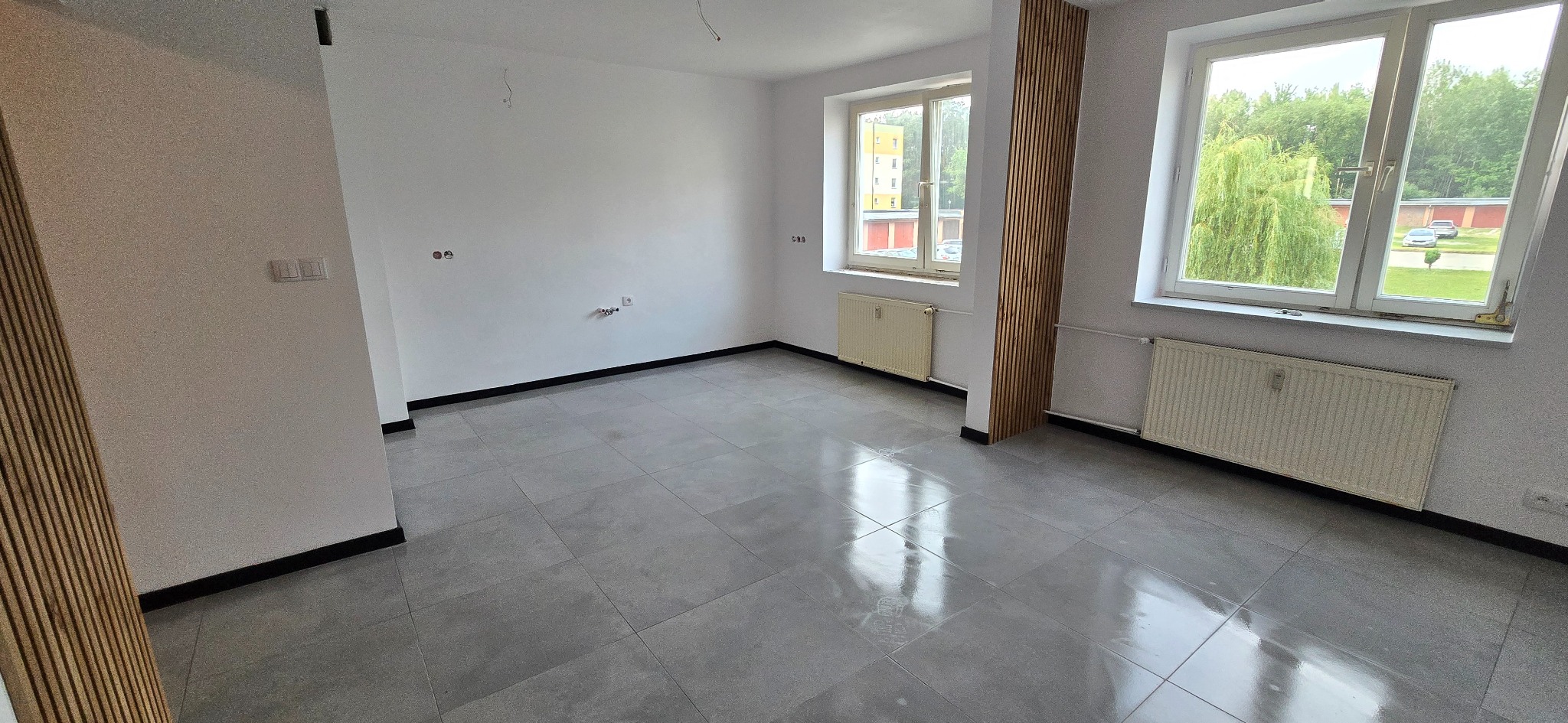 Wykończone wnętrze: szare płytki podłogowe, białe ściany z drewnianymi panelami dekoracyjnymi i grzejnikami pod oknami. Widok na zieleń za oknem.