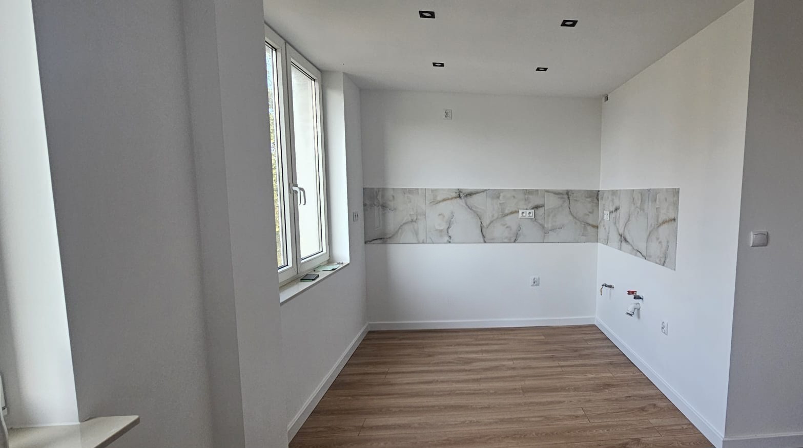 Jasna kuchnia po remoncie: marmurowe płytki nad blatem, drewniana podłoga, białe ściany i okno z widokiem na zieleń. Minimalistyczne oświetlenie sufitowe.