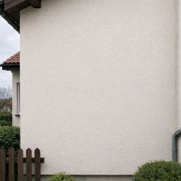 Myjemydomy - Jasna elewacja domu z widocznym fragmentem dachu z brązową dachówką, rynną i drewnianym płotkiem. Krzewy przy ścianie. Ujęcie z boku budynku.