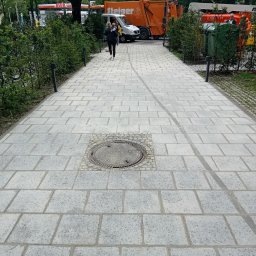 TOMASZ CHRZANOWSKI - Szary chodnik z kostki brukowej w parku, z centralnie umieszczoną studzienką kanalizacyjną otoczoną mniejszą kostką. W tle zieleń, pojazdy komunalne i przechodząca osoba.