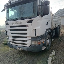 TOMASZ CHRZANOWSKI - Biała ciężarówka Scania R420 z pomarańczowym światłem ostrzegawczym na dachu, zaparkowana na ziemi z kostkami obok. Widok z przodu, pochmurne niebo w tle.