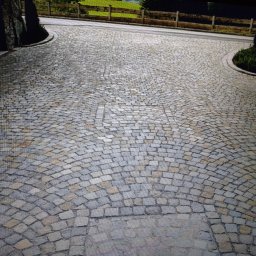 Tomasz Chrzanowski - Ułożona kostka granitowa w łukowaty wzór na podjeździe w Kamienniku. Widoczne kamienne obrzeża i fragment drewnianego płotu w tle. Precyzyjne wykonanie.
