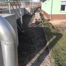 TOMASZ CHRZANOWSKI - Teren po wykopie pod instalacje, widoczne rury, paleta z kostką brukową i fragment budynku z rynną. Przygotowanie do dalszych prac.