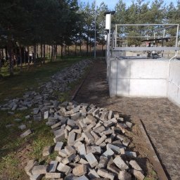 Tomasz Chrzanowski - Sterta rozbiórkowej kostki brukowej przy betonowej konstrukcji na tle drzew i błękitnego nieba. Widoczny fragment ścieżki i trawy.