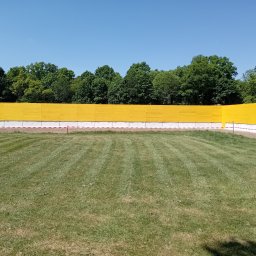 TOMASZ CHRZANOWSKI - Żółte ogrodzenie terenu budowy z widocznymi drzewami w tle i równo przystrzyżonym trawnikiem na pierwszym planie, w słoneczny dzień.