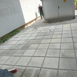 TOMASZ CHRZANOWSKI - Ułożona szara kostka betonowa z drobnym żwirem w spoinach, widok z góry, z boku ściana budynku i szafa elektryczna, w tle osoba w odzieży roboczej.