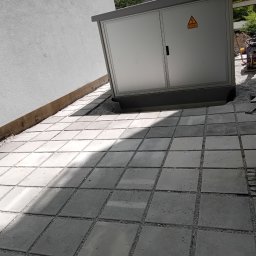 TOMASZ CHRZANOWSKI - Ułożona, szara kostka brukowa kwadratowa z drobnym żwirem w spoinach, obok ściany budynku i skrzynki elektrycznej. W tle ubijarka i kostka granitowa.