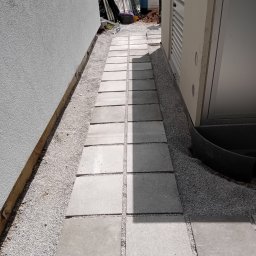 TOMASZ CHRZANOWSKI - Ścieżka z kwadratowych płyt betonowych ułożona wzdłuż budynku, z drobnym żwirem wypełniającym przestrzenie. Widok z góry, oświetlenie słoneczne.