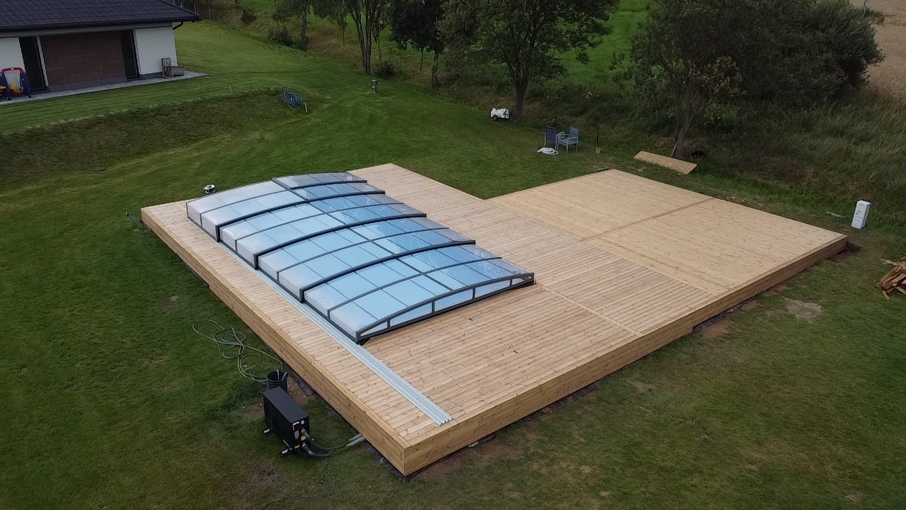 Jasny, drewniany taras z zadaszeniem basenowym na zielonym trawniku. Widok z góry ukazuje regularny układ desek i nowoczesną konstrukcję zadaszenia.