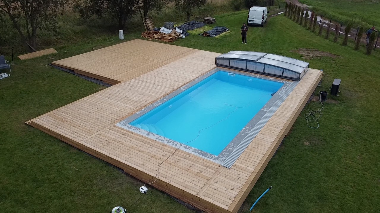 Nowoczesny basen otoczony drewnianym tarasem z jasnego drewna, widok z góry. Część tarasu w trakcie budowy, w tle trawnik i zabudowania gospodarcze.