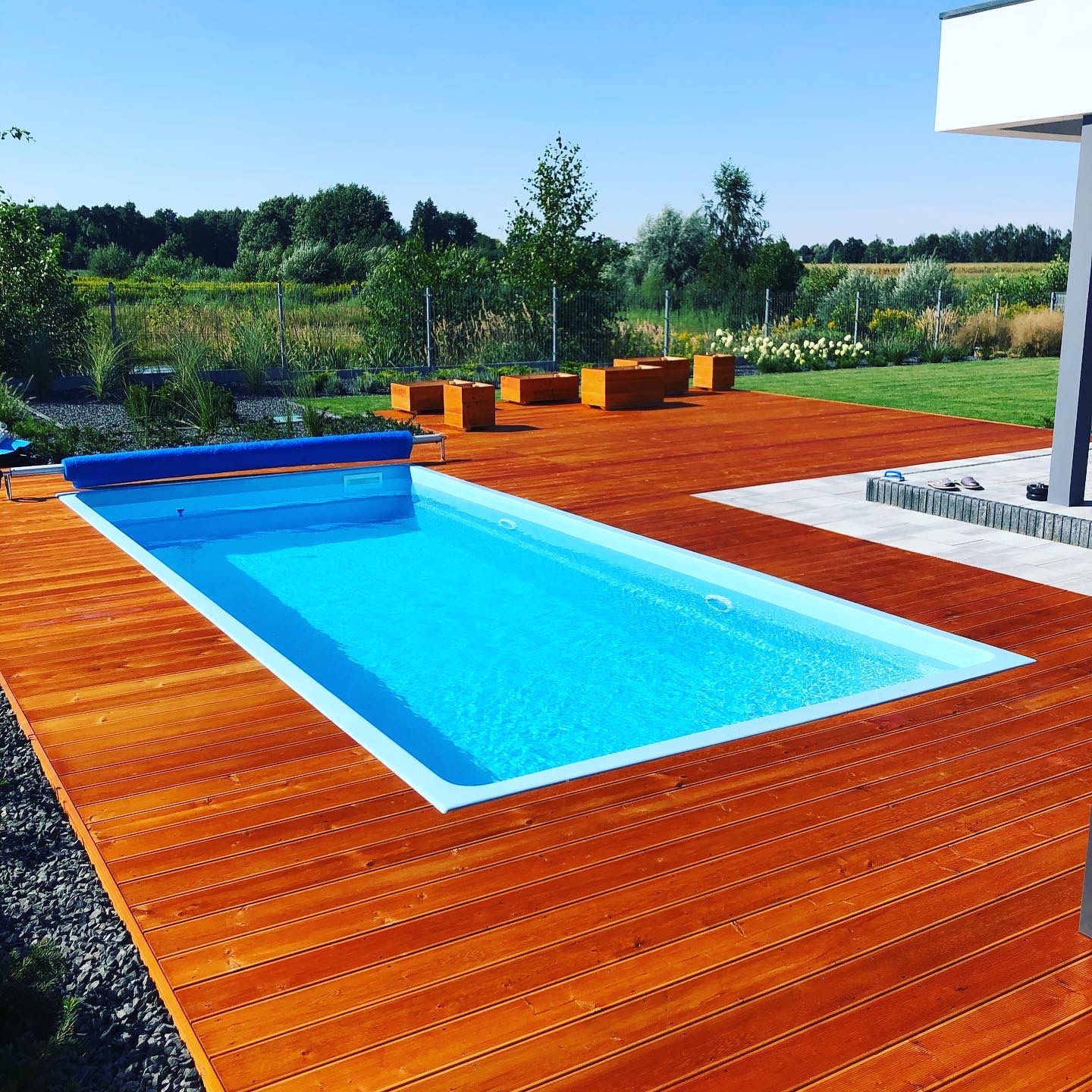 Drewniany taras otaczający basen z błękitną wodą, w tle zielony trawnik i drzewa. Widoczne drewniane donice i fragment nowoczesnego domu.