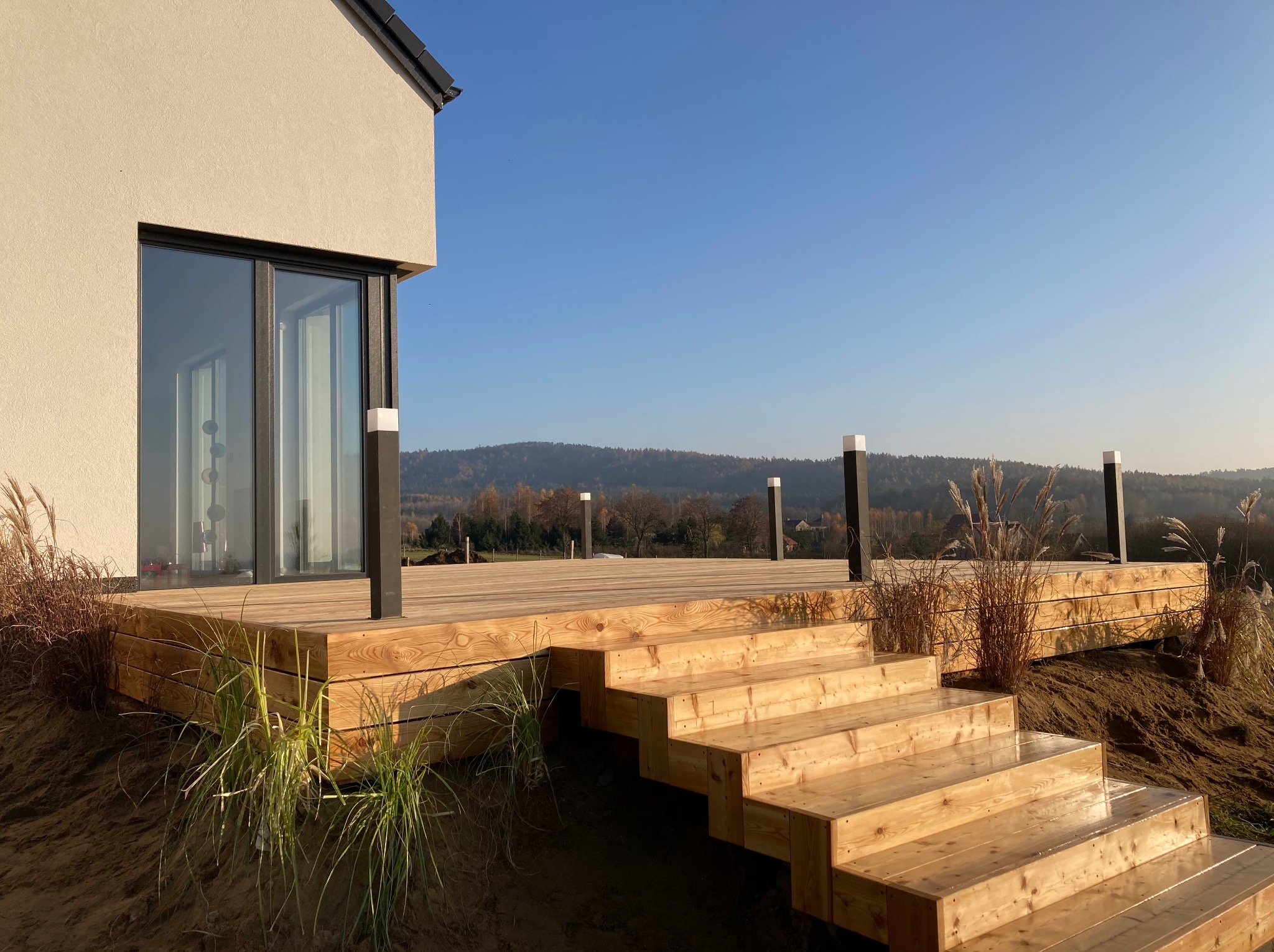 Nowoczesny taras z drewna z niskimi schodami, oświetleniem punktowym i ozdobnymi trawami na tle błękitnego nieba i zalesionych wzgórz. Jasne drewno, minimalistyczny design.