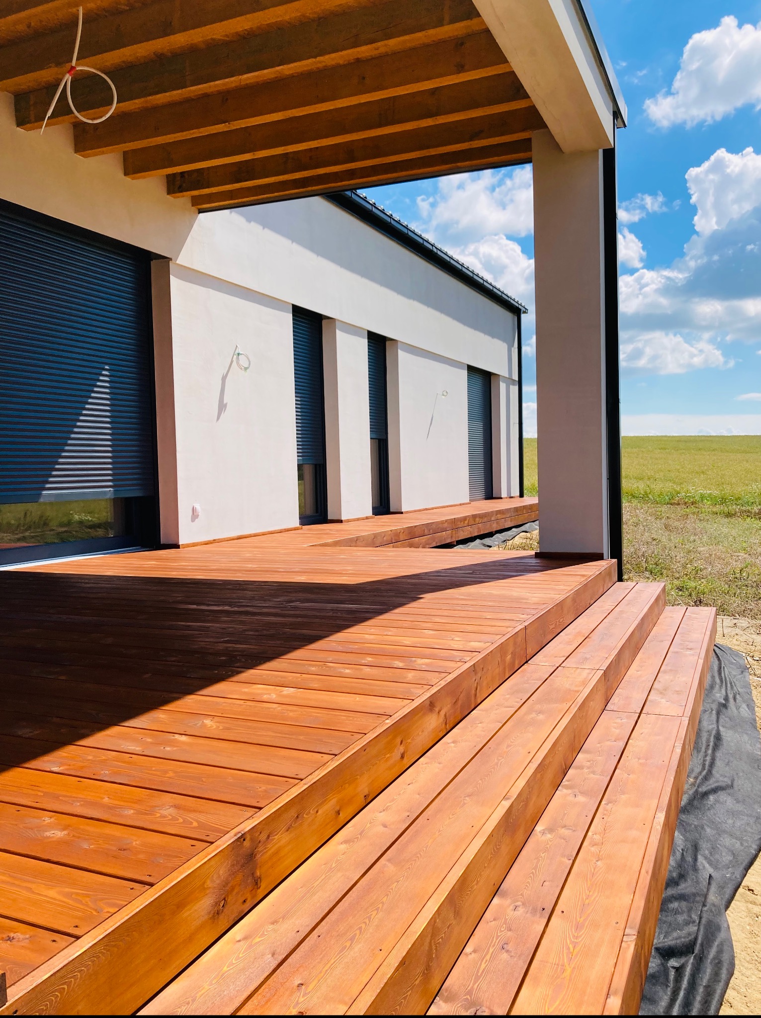 Nowoczesny taras z drewna egzotycznego przy białym domu z roletami zewnętrznymi. Widok na zielone pole i błękitne niebo z chmurami. Jasne, naturalne światło.