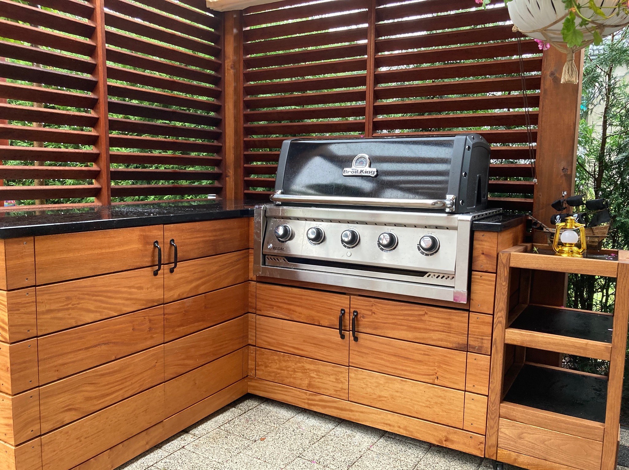 Drewniana zabudowa grilla na tarasie z blatem z czarnego kamienia i ażurową ścianką z drewna. Grill marki Broil King. Widok na zieleń w tle.