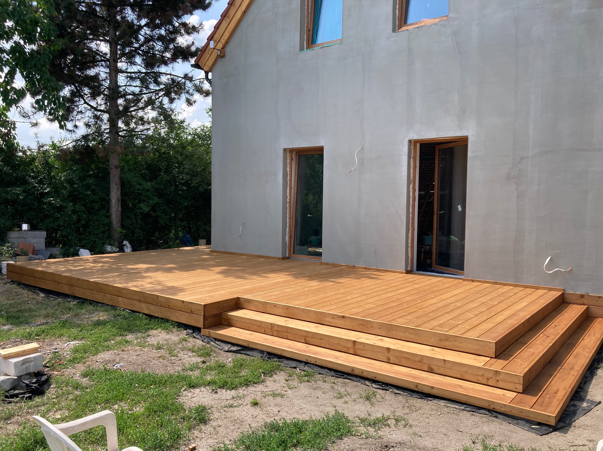 Nowy, drewniany taras z trzema schodkami, przylegający do surowego budynku w trakcie budowy. Widoczne świeże drewno i elementy wykończeniowe.