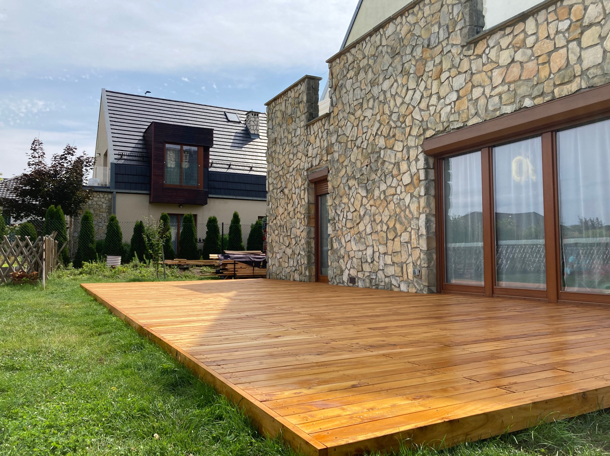Nowy, drewniany taras przydomowy z desek w ciepłym odcieniu brązu, na tle domu z kamienną elewacją i zielonego trawnika. Widok z poziomu trawy, słoneczny dzień.