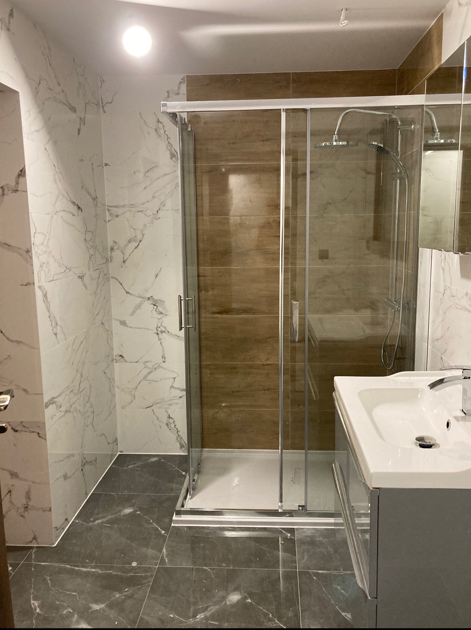 Nowoczesna łazienka z kabiną prysznicową typu walk-in, marmurowymi płytkami na ścianach i szarą podłogą. Umywalka z szafką w kolorze szarym. Widoczne detale wykończenia.