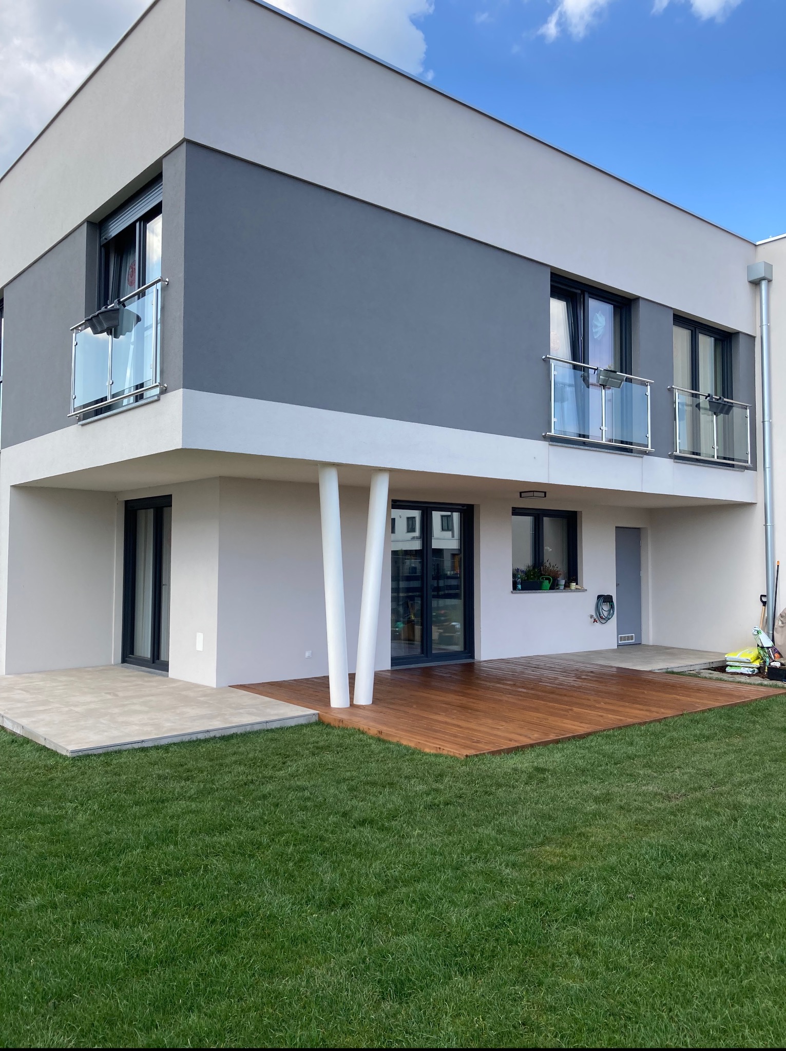 Nowoczesny dom z drewnianym tarasem, widok z ogrodu. Szklane balustrady balkonowe, minimalistyczna architektura, zadbany trawnik. Elementy wykończeniowe w odcieniach szarości i bieli.