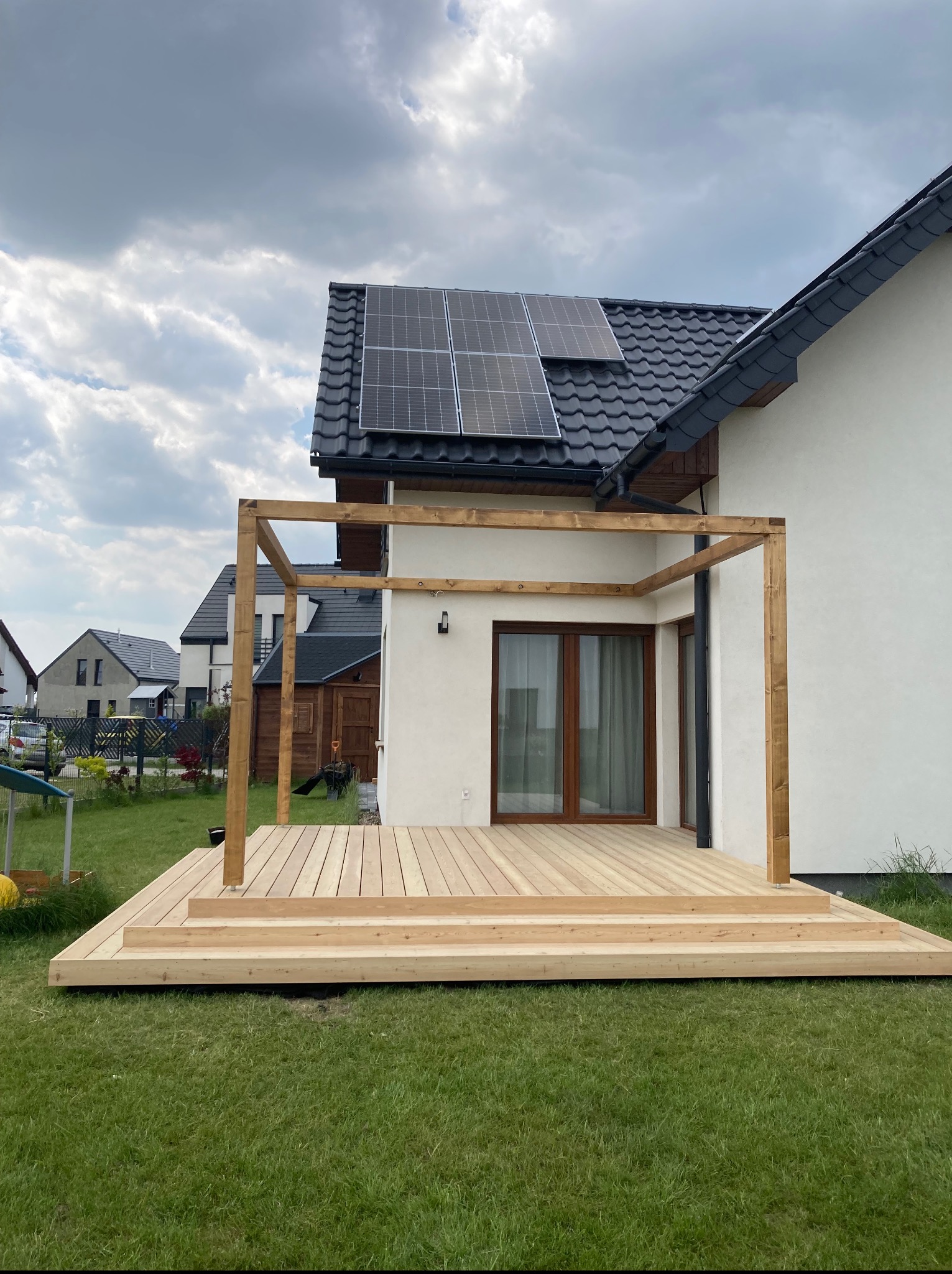 Nowoczesny taras drewniany z surowego drewna z elementami pergoli, na tle domu z panelami słonecznymi i zielonego trawnika. Widok z poziomu trawy.