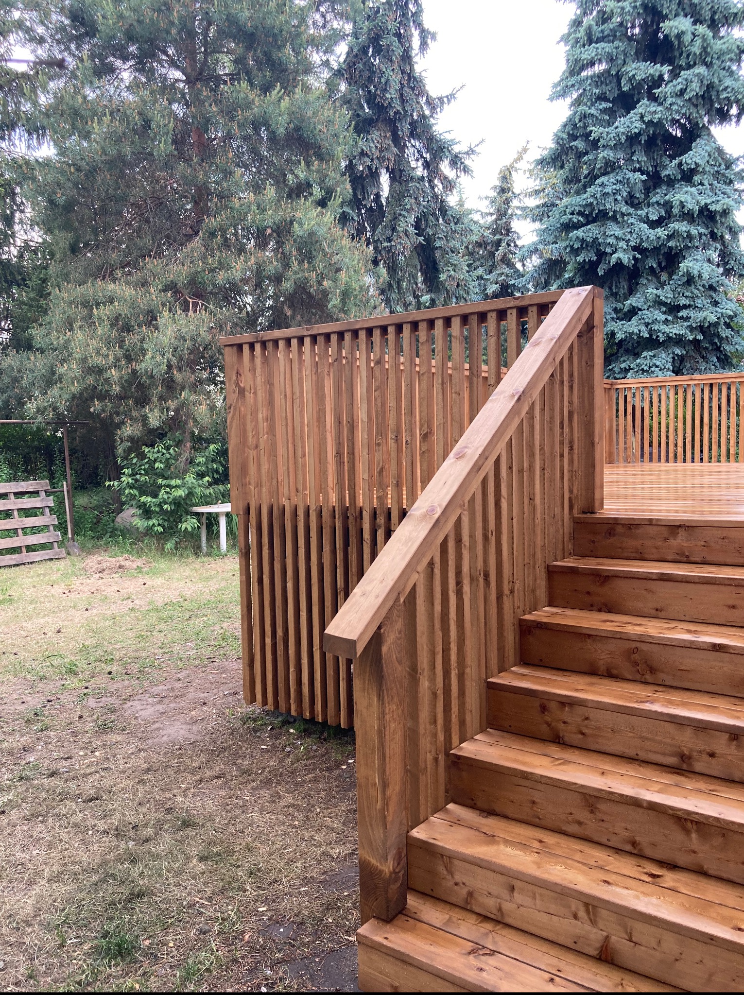 Drewniany taras z balustradą i schodami, otoczony zielenią. Widoczna drewniana konstrukcja osłonowa. Ujęcie z dołu, podkreślające solidność wykonania.