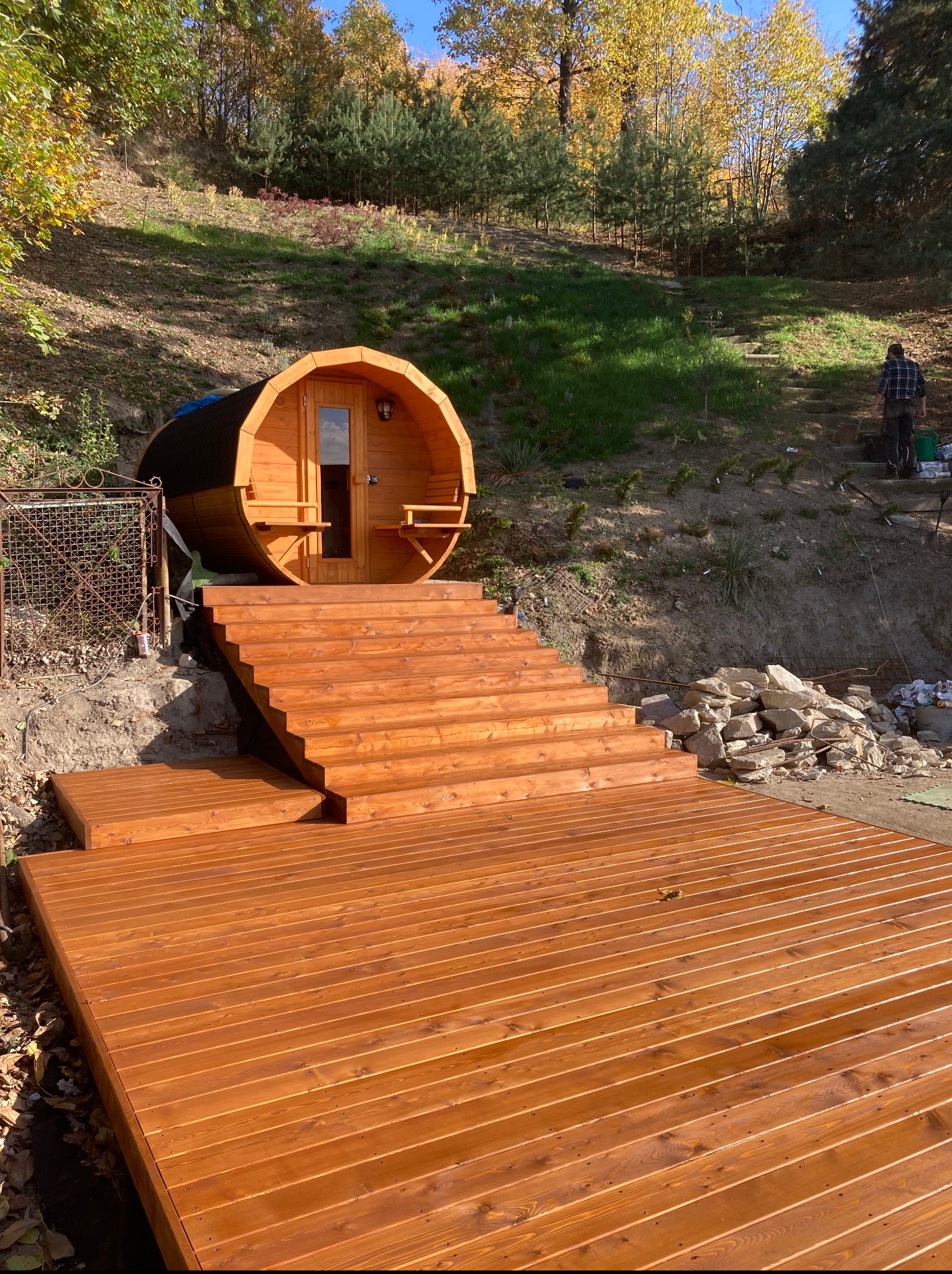 Drewniany taras z beczką sauną na wzgórzu, schody prowadzą do wejścia. W tle drzewa i osoba pracująca na zboczu. Ciepłe światło słoneczne podkreśla fakturę drewna.