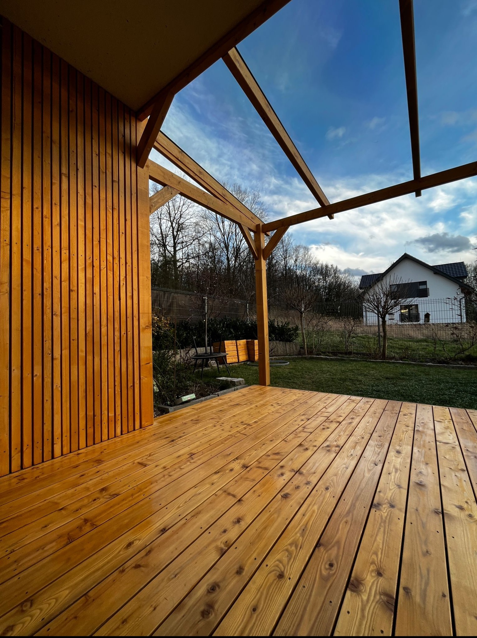 Drewniany taras z pergolą w ogrodzie, widok na dom. Deski tarasowe w ciepłym odcieniu brązu, pionowe deski na ścianie. Błękitne niebo z chmurami w tle.