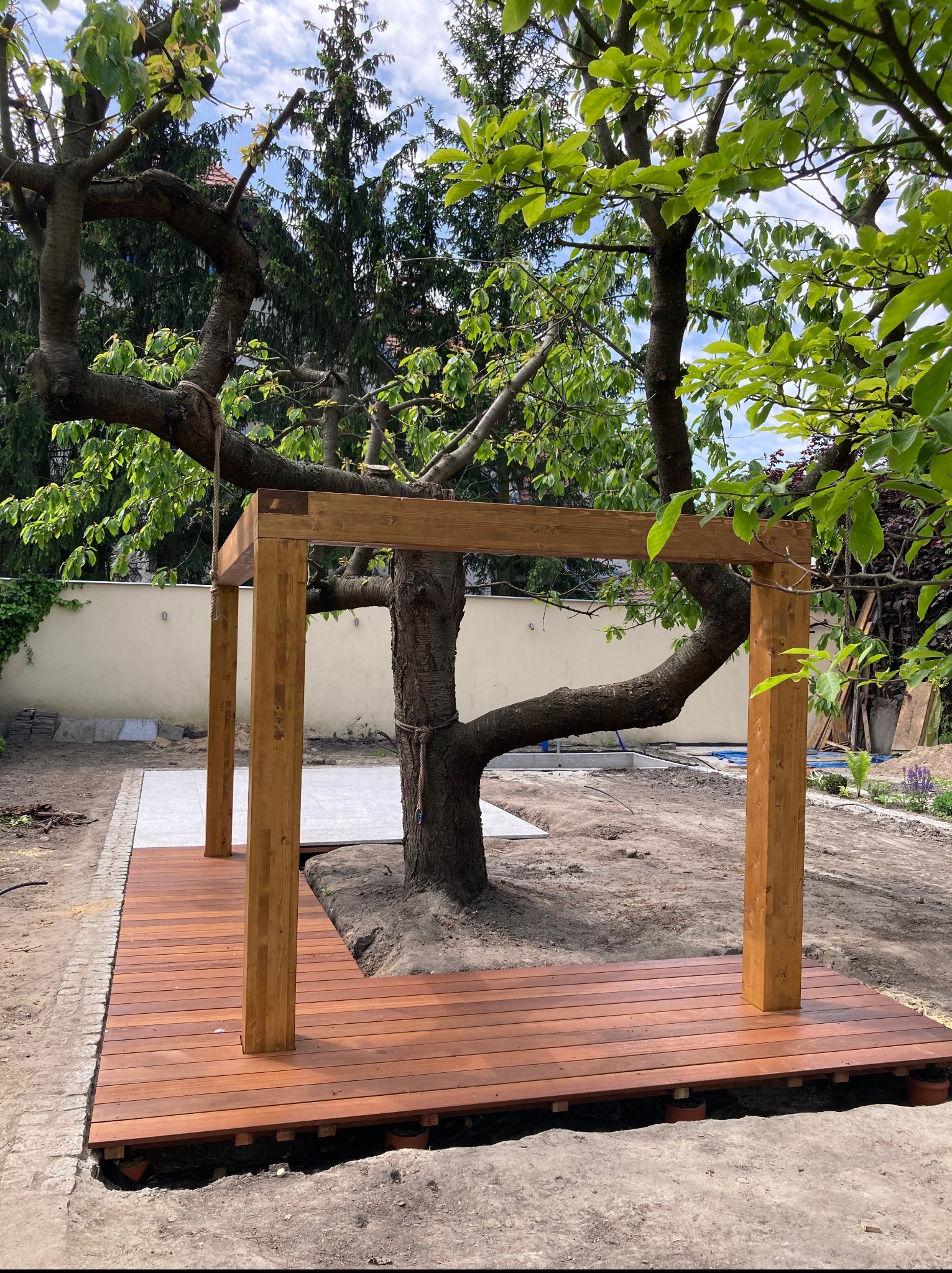 Drewniany taras z konstrukcją pergoli wokół drzewa, w trakcie budowy. Widoczne deski tarasowe i drewniane słupy. Tło stanowi ogród z zielenią i jasnym murem.