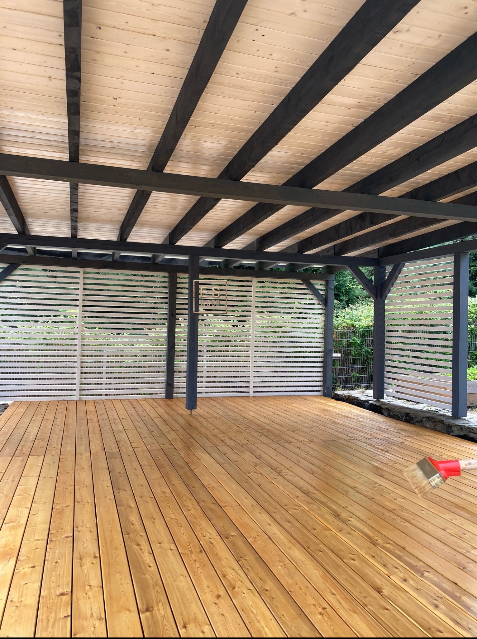 Zadaszony taras z drewnianą podłogą, pomalowaną na złoty kolor. Widoczne białe, poziome panele boczne i ciemne belki konstrukcyjne. Pędzel do malowania w prawym dolnym rogu.