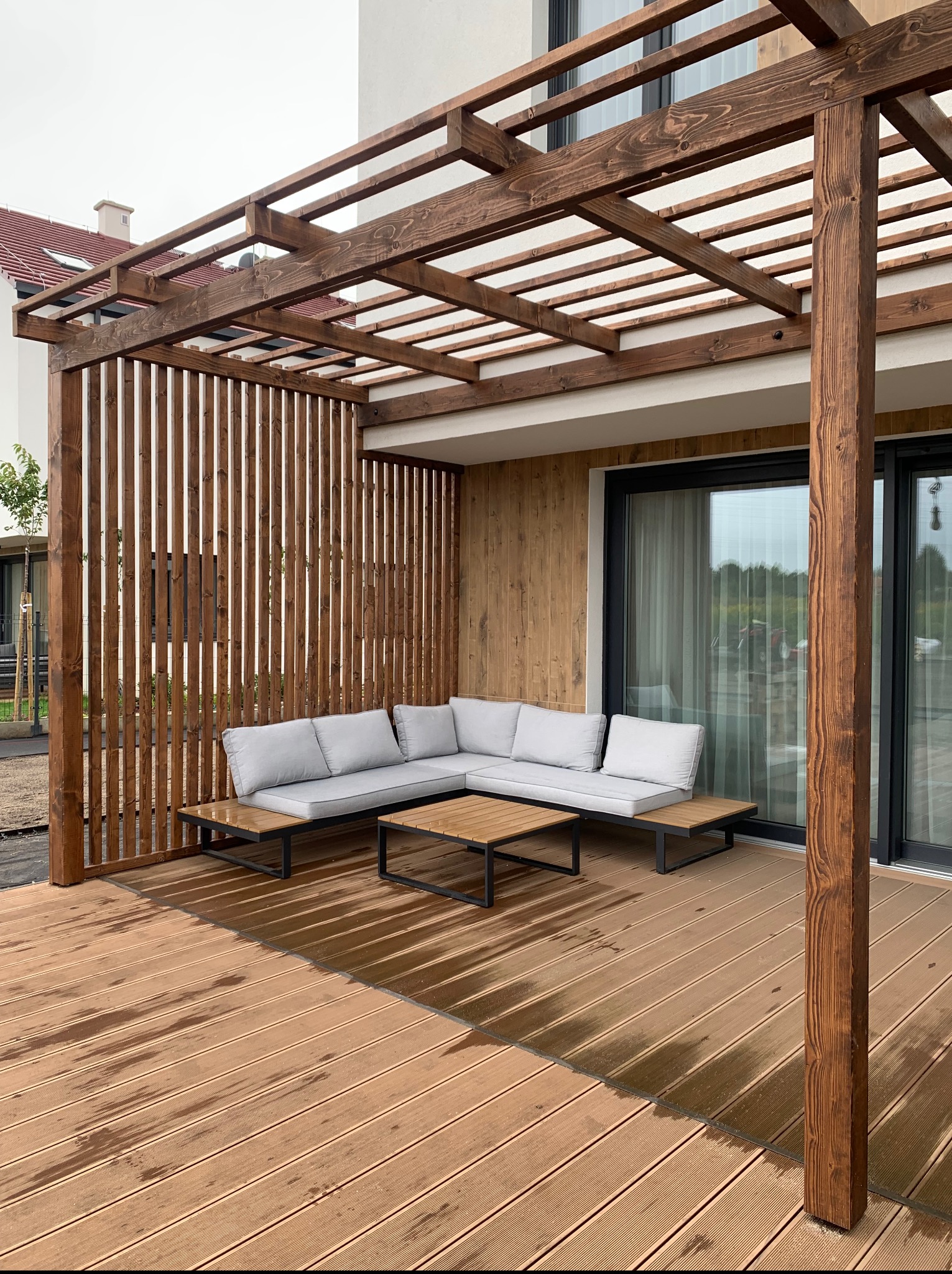 Nowoczesny taras drewniany z pergolą i szarą sofą narożną. Widoczne deski tarasowe z wyraźnym usłojeniem i drewniana zabudowa pionowa. Całość w neutralnej kolorystyce.