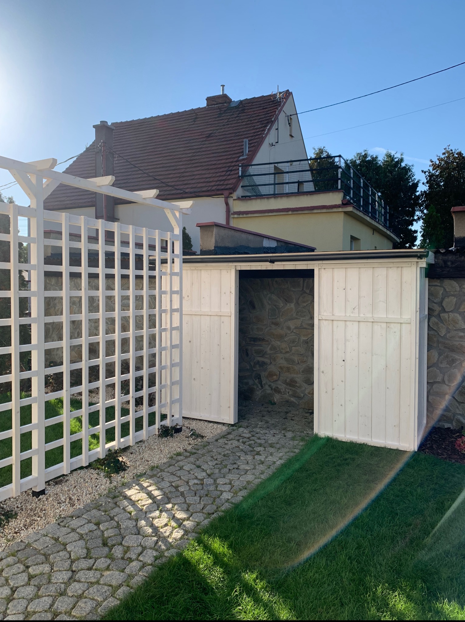 Biała pergola z kratką i drewniana szopa z otwartymi drzwiami na kamiennym podłożu, w tle dom z czerwoną dachówką i zielony trawnik.