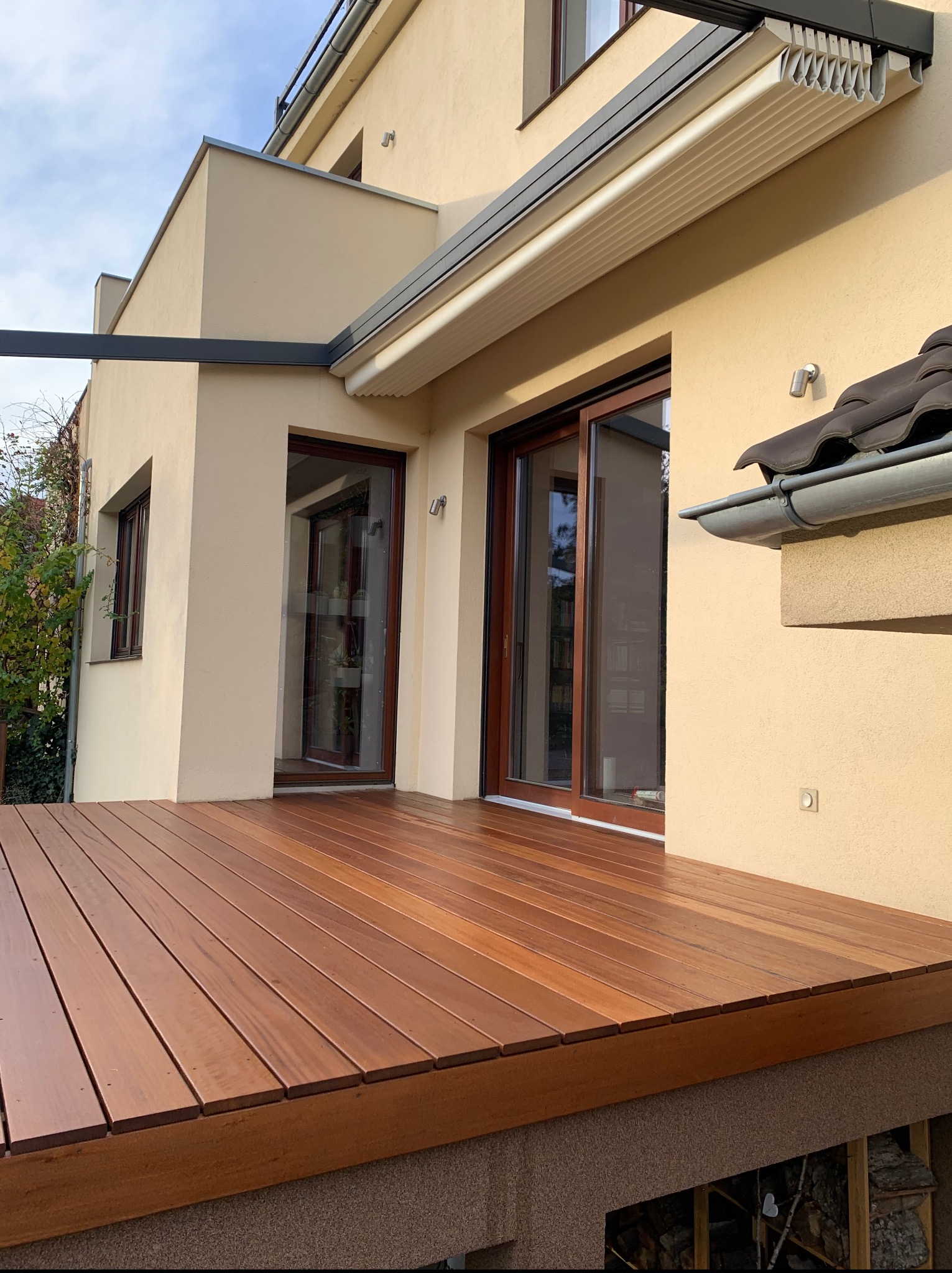 Nowoczesny taras z drewna egzotycznego przy beżowym domu. Widoczne przesuwne drzwi tarasowe, pergola i starannie ułożone deski tarasowe z widoczną strukturą drewna.